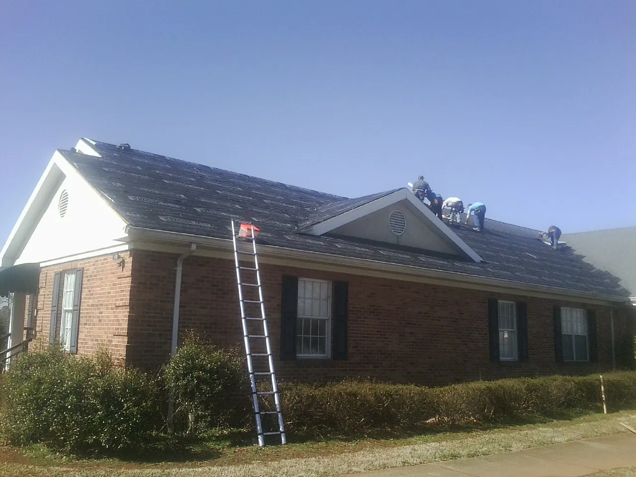 Crew tearing shingles off a brick ranch roof with a ladder set — Emergency Roof Repair work by our crew in Manakin Sabot