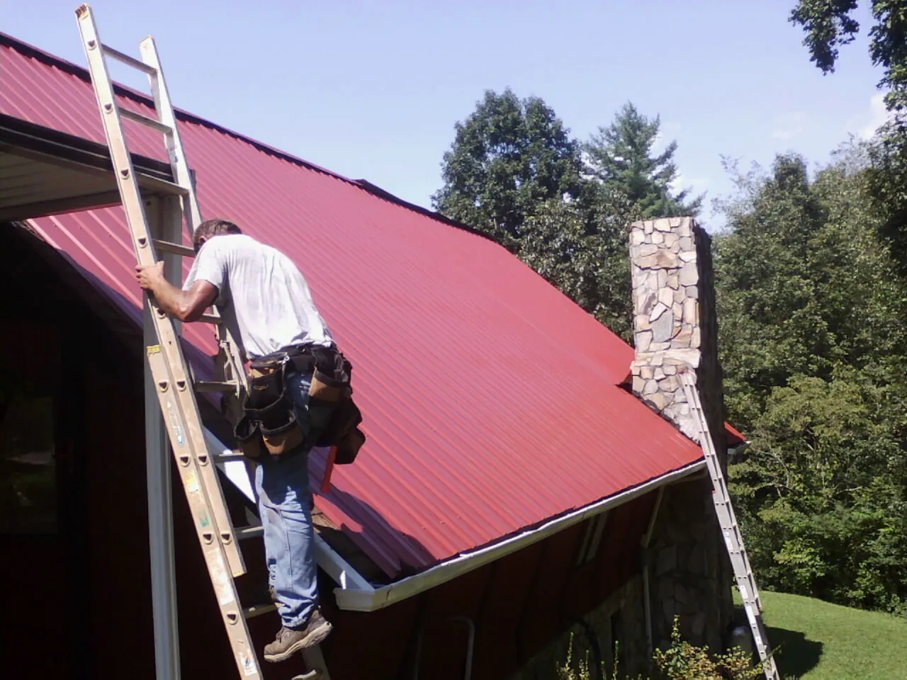 Roofer on a ladder beside a red metal roof and stone chimney — Soffit & Fascia Repair by Roofing Brothers in Manakin Sabot